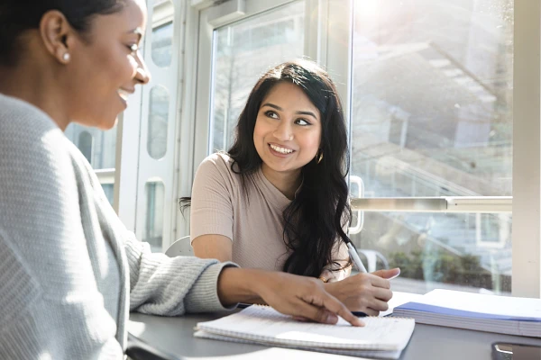 Two women are sitting at a table, smiling and talking, with notebooks in front of them.