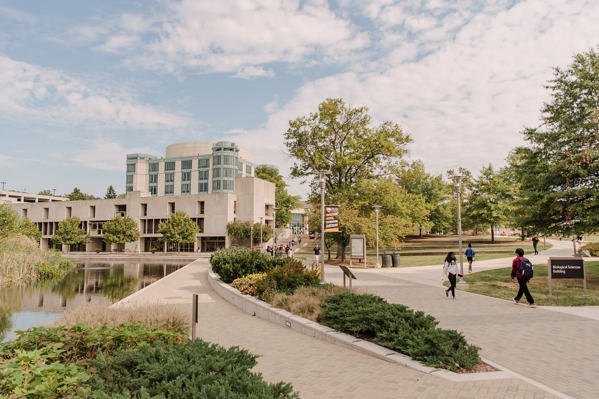 A modern campus building surrounded by trees and pathways with people walking nearby.